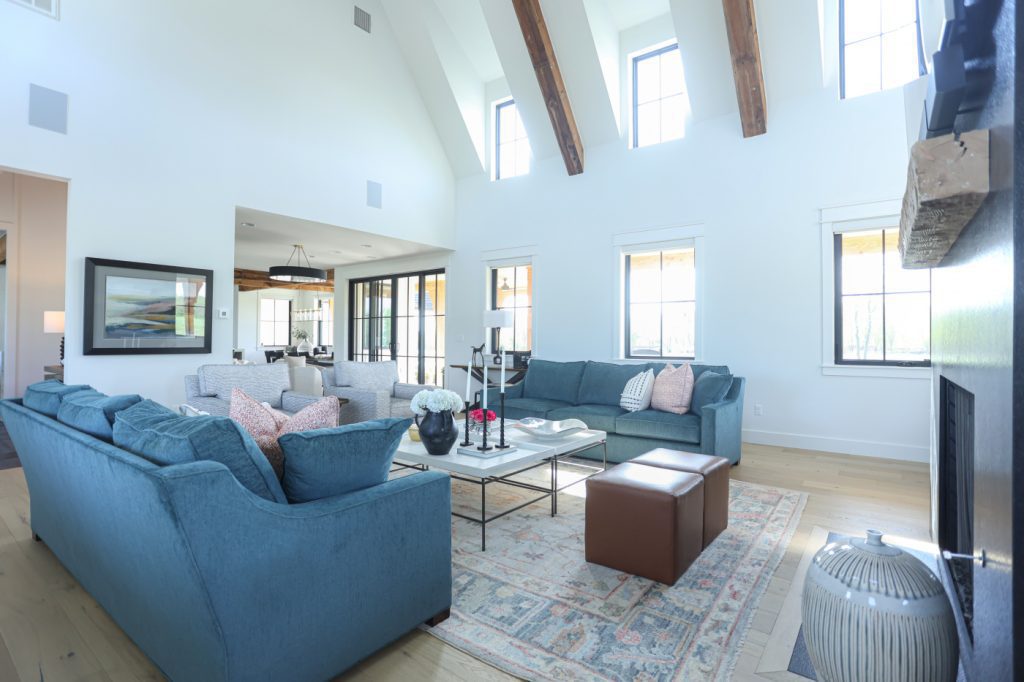 View of a spacious living room from the black granite fireplace wall, showcasing tall windows, reclaimed wood ceiling beams, layered seating with colorful sofas and patterned accents, and a one-of-a-kind rug that adds warmth to the neutral black-and-white backdrop.