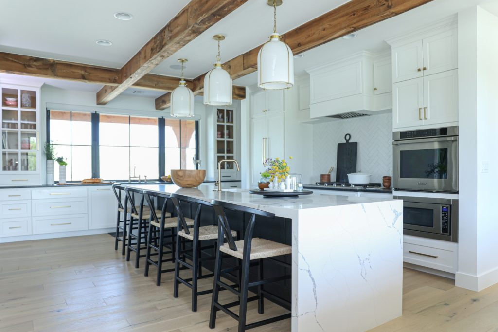 Luxury kitchen by Kirkendall Design in Tulsa, OK showcasing reclaimed wood beams, a kitchen island with Bianco Statuario Quartz, Hudson Valley pendant lighting, large windows overlooking the front yard, and custom cabinetry.