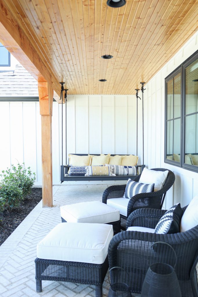Luxury interior design by Kirkendall Design in Tulsa, OK showcasing the exterior of a new construction home with a wrap-around porch featuring multiple outdoor gathering areas, including a fire pit, daybed swing, outdoor fireplace, dining and grilling area, lounge off the primary suite, and swings from cedar beams for children.