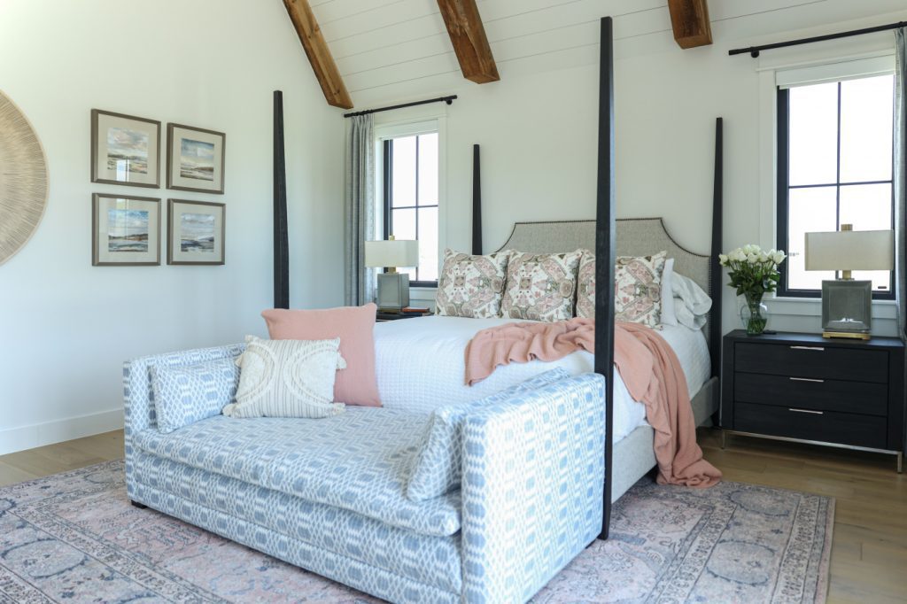 Luxury interior design by Kirkendall Design in Tulsa, OK showing a primary bedroom with an oversized four-poster bed, seating area, remote-controlled shades, blackout curtains, and patterned accents in blue, white, and peachy pink.