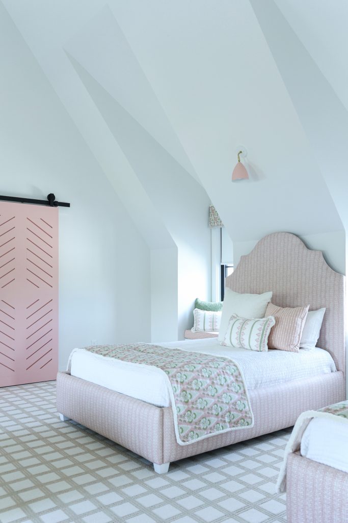 Luxury interior design by Kirkendall Design in Tulsa, OK showing a girls’ bunk room with four custom beds, individual window seats at dormer windows, a neutral windowpane-patterned carpet, and rosy pink accents in bedding and sconce shades.