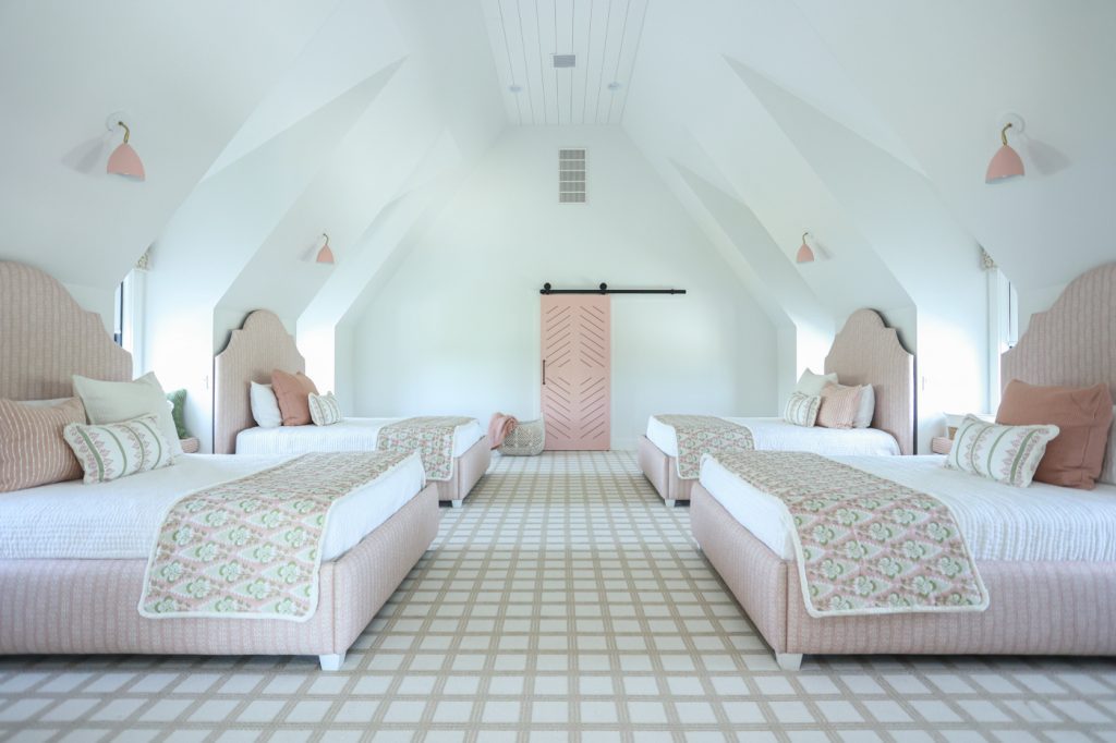 Luxury interior design by Kirkendall Design in Tulsa, OK showing a girls’ bunk room with four custom beds, individual window seats at dormer windows, a neutral windowpane-patterned carpet, and rosy pink accents in bedding and sconce shades.