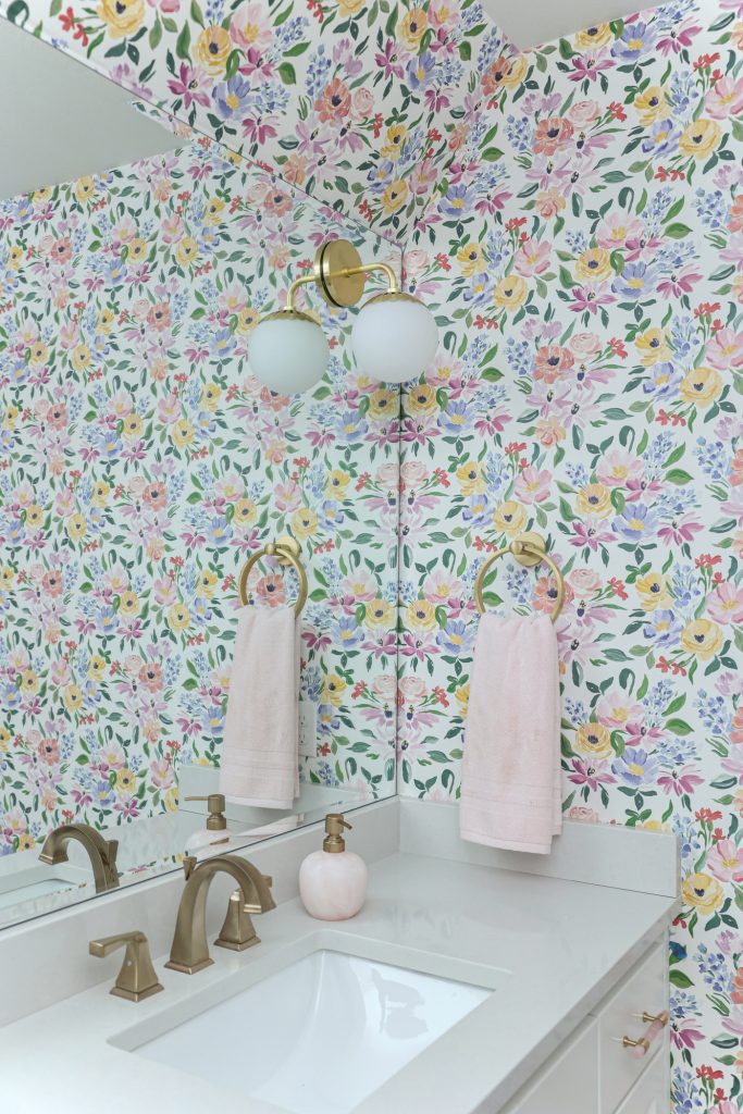 Luxury interior design by Kirkendall Design in Tulsa, OK featuring a girls’ bathroom with bright floral wallpaper, round wall sconces, black-and-white starburst tile, and a white vanity.