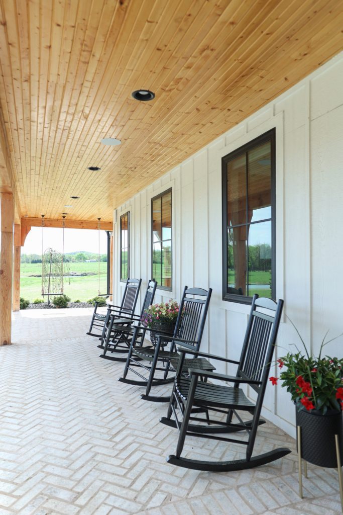 Luxury interior design by Kirkendall Design in Tulsa, OK showcasing the exterior of a new construction home with a wrap-around porch featuring multiple outdoor gathering areas—including a fire pit, daybed swing, outdoor fireplace, dining and grilling area, lounge off the primary suite, and swings from cedar beams—with brick flooring laid in a herringbone pattern.