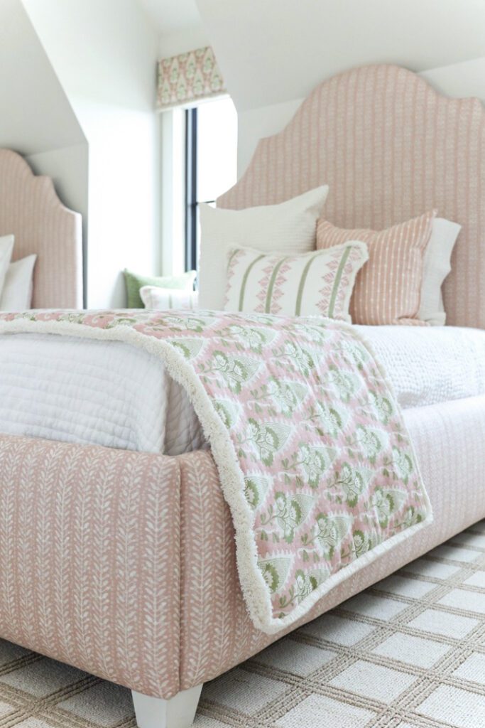 Luxury interior design by Kirkendall Design in Tulsa, OK showing a girls’ bunk room with four custom beds, individual window seats at dormer windows, a neutral windowpane-patterned carpet, and rosy pink accents in bedding and sconce shades.