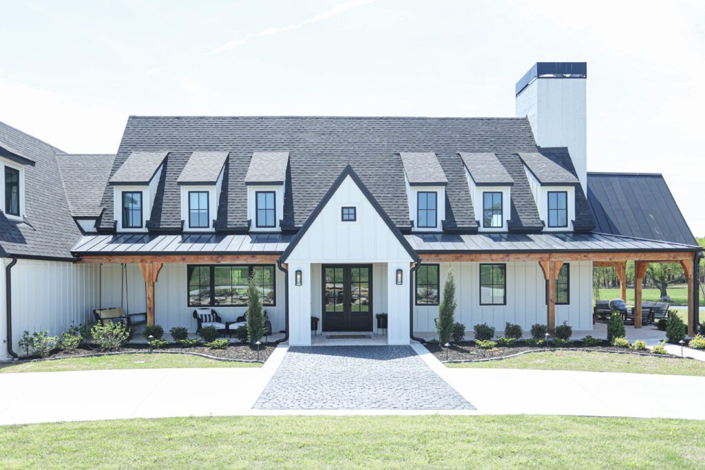 Exterior view of a luxury new-construction ranch home near Tulsa, designed by Kirkendall Design. The house features cedar posts, black-frame windows, metal roofing, and a wrap-around porch with herringbone brick flooring—showcasing modern farmhouse sophistication and inviting outdoor living spaces.