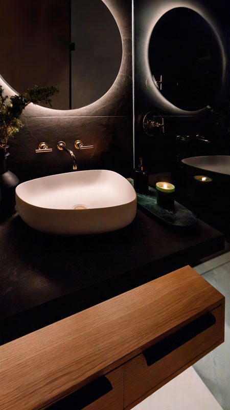 NKBA 2025 award-winning bathroom with a moody aesthetic featuring black tile backsplash, round backlit mirror and vessel sink.