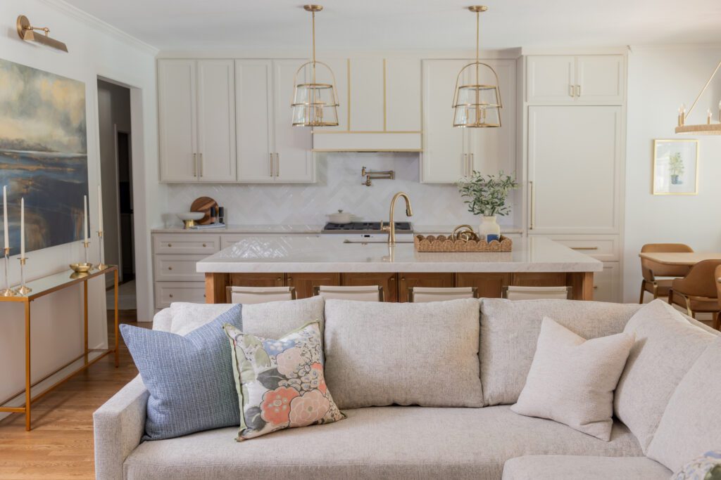 Luxury interior design by Kirkendall Design in Tulsa, OK featuring a kitchen renovation with an open layout, central island in stained weathered oak, soft neutral cabinetry, quartz countertops, classic ceramic tile backsplash, integrated appliances, and a custom hood with satin brass detailing.