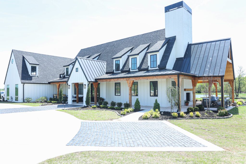 Exterior view of a luxury new-construction ranch home near Tulsa, designed by Kirkendall Design. The house features cedar posts, black-frame windows, metal roofing, and a wrap-around porch with herringbone brick flooring—showcasing modern farmhouse sophistication and inviting outdoor living spaces.