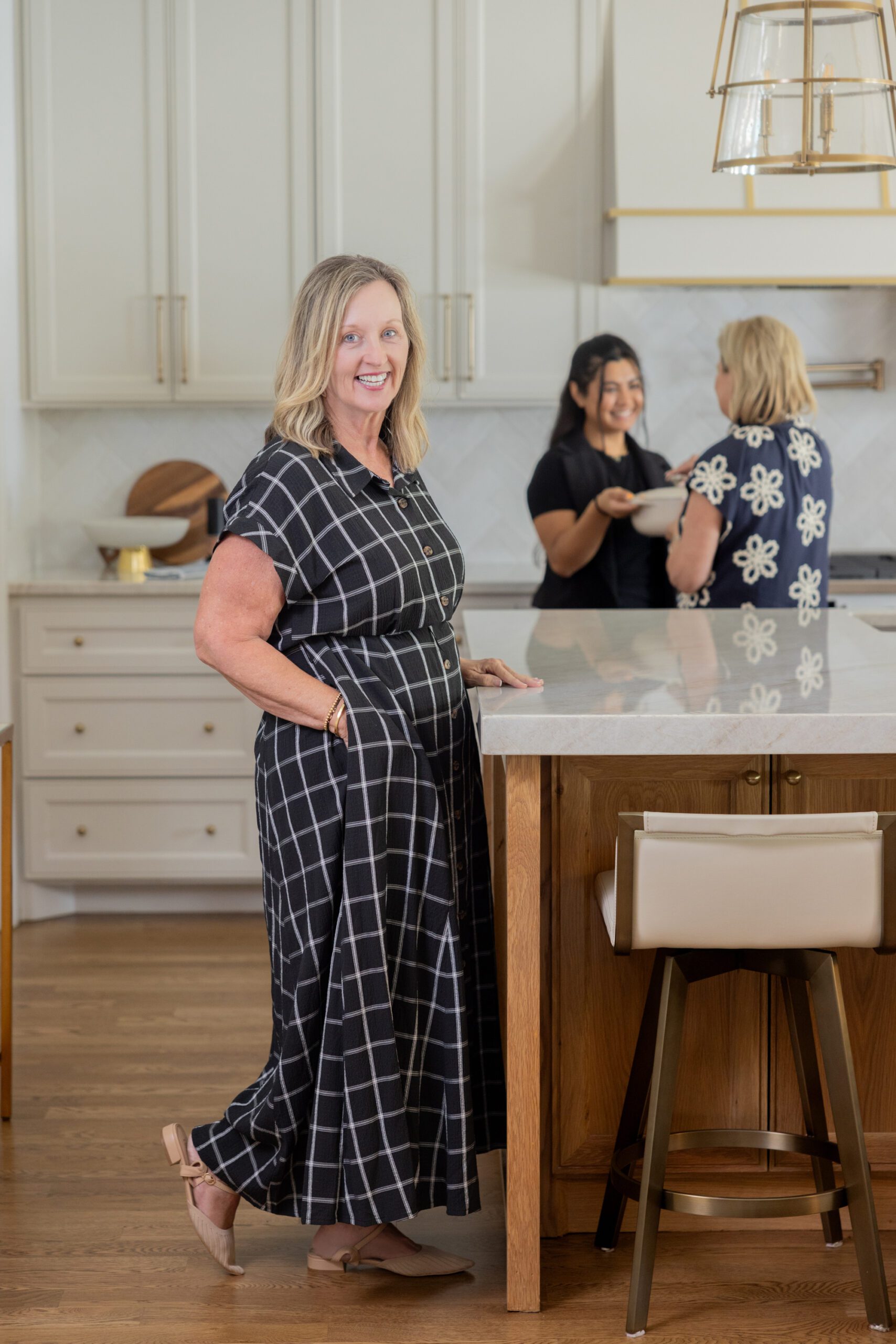 Luxury interior design by Kirkendall Design in Tulsa, OK featuring a kitchen renovation with an open layout, stained weathered oak island, neutral cabinetry, quartz countertops, classic ceramic tile backsplash, integrated appliances, and custom hood with satin brass detailing, with designer and owner Julia Kirkendall pictured in the space.