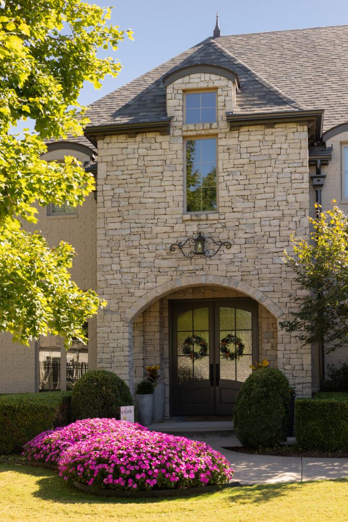 Tuscan-Tudor style home in Tulsa, transformed with exterior stone limewash paint. The exterior refresh also included newly painted trim, a darker roof, an updated garage door, and the removal of shutters